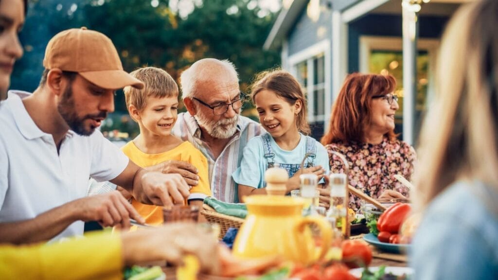 A multigenerational family of a man, a boy, an older adult man, a girl, and an older adult woman sits at a table outside. There is food on the table.