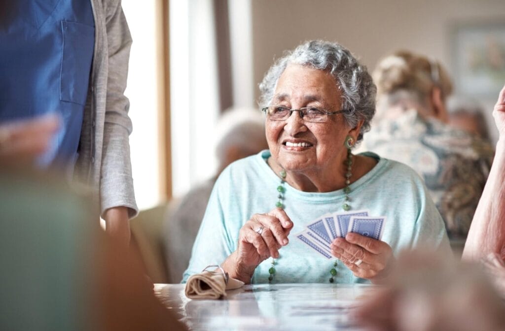 An older adult woman sits at a table, holding a hand of playing cards. Just out of frame is a caregiver. There are other older adults at her table and at a table behind her, all out of focus.