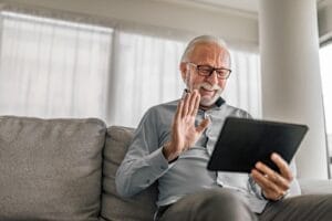 An older adult man is sitting on his couch, holding a tablet device, and waving at someone he is video chatting with.