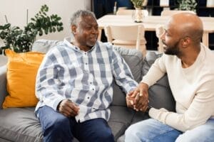 A man sits on a couch with his older adult father, holding his hand.